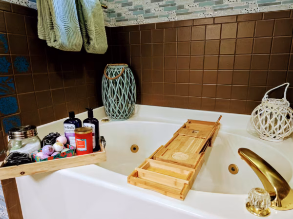 A corner bathtub features a wooden caddy resting across its width and a wooden tray on the rim holding bath bombs, bottles of soap, and a red candle.