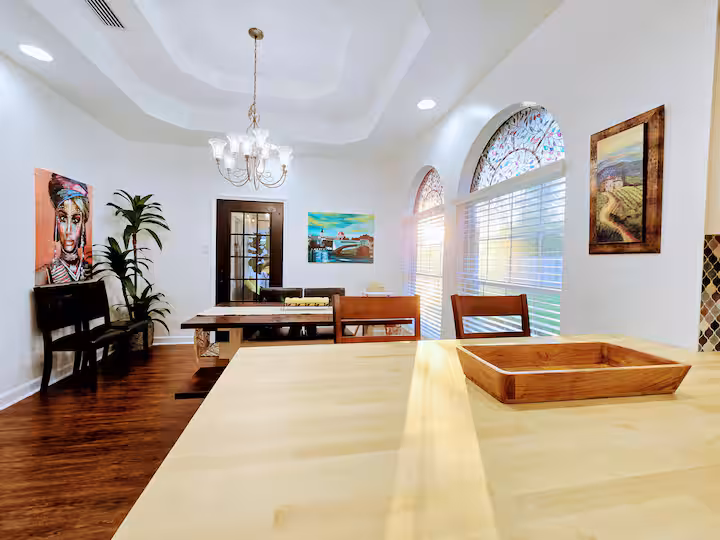 A view from a light wood kitchen island countertop looking across into an open-concept dining area featuring a dark wooden dining table, a traditional chandelier suspended from a tray ceiling, arched windows, and colorful artwork on the white walls.