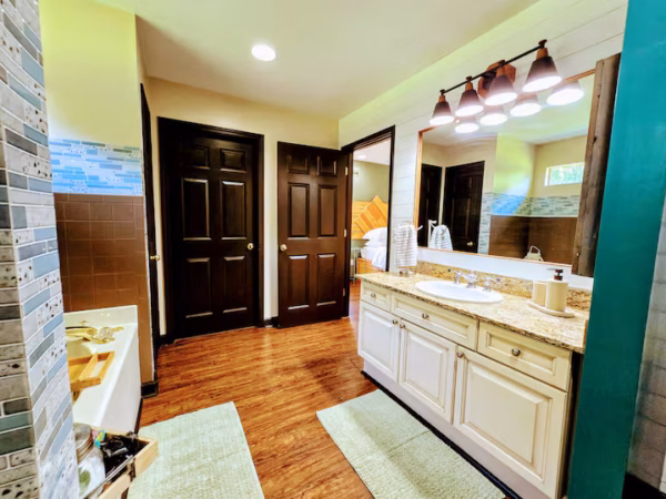 An eye-level view of a bright bathroom featuring dark brown wooden doors, a white double vanity with a marble countertop, and wood-look flooring.